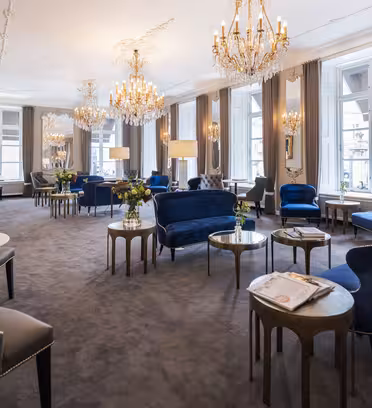 Elegant hotellsalong med overdådige lysekroner, store speil og gulv-til-tak-vinduer. Rommet har myke lenestoler og sofaer i blå fløyel, bord med marmorplater og høye stående lamper, som skaper en sofistikert atmosfære.