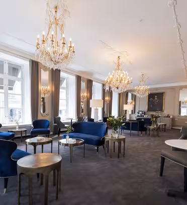 Elegant salong med utsmykkede lysekroner, blå fløyelsstoler og små runde bord på et mykt teppe. Store vinduer med gardiner langs veggen, lyser opp rommet. Dekorative speil og klassiske malerier forsterker den sofistikerte stemningen.