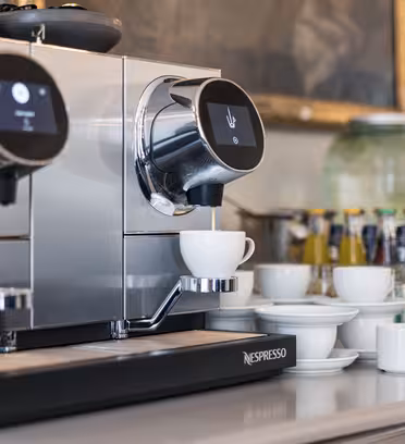 En moderne espressomaskin som brygger kaffe til en hvit kopp. Den har digitale skjermer og er omgitt av ekstra kopper og tallerkener. I bakgrunnen er det ulike drikkeflasker og glasskrukker på en disk.