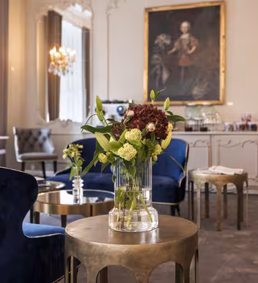 Elegant rom med fløyelsstoler, utsmykkede speil og et stort maleri på veggen. En vase med røde og hvite blomster står på et lite rundt bord i forgrunnen. Lysekroner og store vinduer bidrar til den sofistikerte atmosfæren.