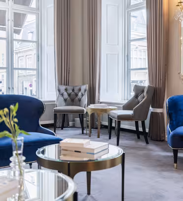 Luksuriøs stue med elegante blå fløyelssofaer og grå stoler, fremhevet av store vinduer med beige gardiner. Et rundt glasssalongbord med bøker og en plante fullfører den sofistikerte stemningen.