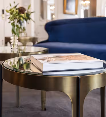 En elegant stue med et rundt salongbord i gull med en blank topp med plass til en stor bok. I bakgrunnen er en blå fløyelssofa og et andre matchende bord med en vase med blomster synlige. Innredningen er elegant og moderne.