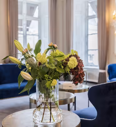 En stilig salong har lenestoler i blå fløyel, elegante lysekroner og store vinduer. I forgrunnen er det et rundt bord med en glassvase med en bukett med grønne og røde blomster.