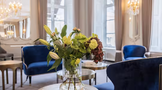 En stilig salong har lenestoler i blå fløyel, elegante lysekroner og store vinduer. I forgrunnen er det et rundt bord med en glassvase med en bukett med grønne og røde blomster.