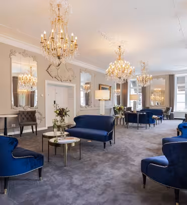 Luksuriøs salong med plysjblå fløyelsstoler og sofaer. Elegante lysekroner henger fra taket, og utsmykkede speil pryder veggene. Innstillingen utstråler raffinement og komfort, med et mykt grått teppe og store vinduer.