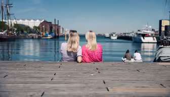 To personer med blondt hår sitter på en trebrygge vendt mot en rolig vannmasse, med båter og et bybilde ved vannet i det fjerne under en klarblå himmel.