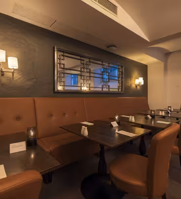 Et svakt opplyst restaurantinteriør med sitteplasser i brunt skinn og mørke trebord. Lampetter på veggene gir dempet belysning. Et stort speil er montert på bakveggen, som gjenspeiler rommets koselige atmosfære.