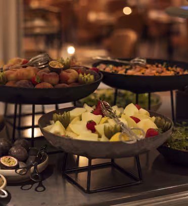 Et buffébord med ulike frukter og salater. Synlige er boller fylt med epler, pærer, kiwi, fersken og blandede grønnsaker, hver med serveringsutstyr. Bakgrunnen viser uskarpe spisestuestoler.