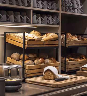 En bakerutstilling med trekasser fylt med ulike typer brød, inkludert rundstykker og brød. Et skjærebrett med brød- og tøyservietter er i forgrunnen, omgitt av en brødrister og andre serveringsartikler. En hylle med krus er over.