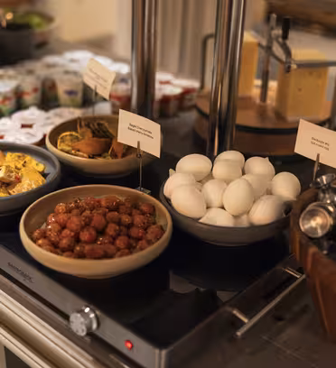 En buffet med ulike retter, inkludert yoghurtbeger, gul eggerøre, kjøttboller i saus, kokte egg og frukt. Osteblokker og stablede metallkopper er også synlige på disken.