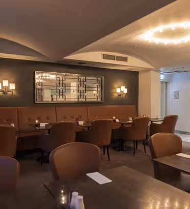 Et koselig restaurantinteriør med brune skinnstoler og mørke trebord. En lang polstret benk langs bakveggen, som har moderne vegglamper og et dekorativt speil. En sirkulær lysekrone henger fra taket.