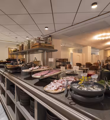 Et elegant bufféoppsett i en restaurant har en rekke kjøttpålegg, oster og grønnsaker på en benkeplate. Spiseplassen i bakgrunnen har pent arrangerte bord og stoler under dempet belysning. Stemningen er varm og innbydende.