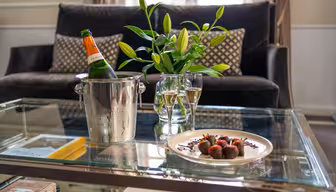 Et glassbord har en tallerken med sjokoladedekkede jordbær, to glass champagne, en isbøtte med en flaske og en vase med liljer. En sofa med mønstrede puter står i bakgrunnen.