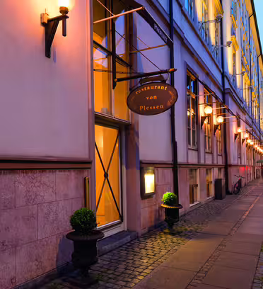 En gatevisning av en bygningsfasade med lysende vegglamper og et skilt med "restaurant von Plessen." Bygningen har store vinduer, et brosteinsbelagt fortau og to små pottebusker. En sykkel står mot veggen.