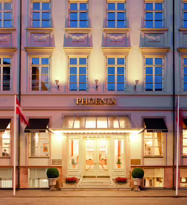 Bildet viser det elegante eksteriøret til Phoenix Hotel, med en klassisk fasade med flere rader med opplyste vinduer. Inngangen er storslått, med to flagg som flankerer døråpningen og potteplanter ved trappen.