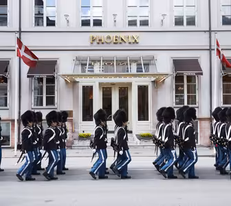 En gruppe uniformerte vakter i svarte hatter og blå bukser marsjerer unisont utenfor Phoenix-bygningen. To flagg med røde og hvite farger vises på hver side av inngangen.