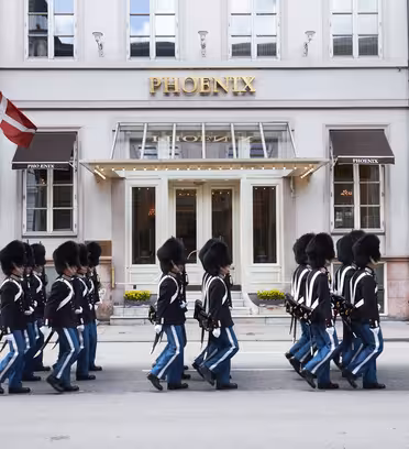 En gruppe uniformerte vakter i svarte hatter og blå bukser marsjerer unisont utenfor Phoenix-bygningen. To flagg med røde og hvite farger vises på hver side av inngangen.
