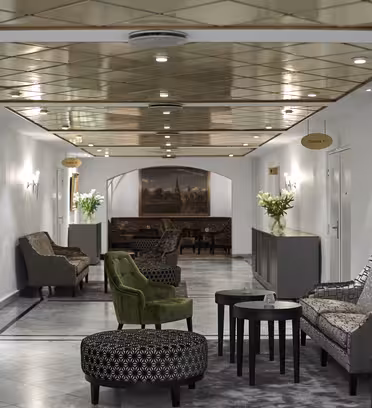 En hotellobby har lenestoler og mønstrede ottomaner langs et marmorgulv. Taket er gullbelagt med innfelt belysning. Veggene er utsmykket med innrammede malerier og vaser med hvite blomster på bordene gir et snev av eleganse.