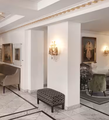 Hotellobby med elegant innredning, med marmorgulv, beige vegger og klassiske lysekroner. Sitteområdet inkluderer en grønn stol, lilla stol og en sofa. Malerier pryder veggene. En trapp med et gyllent rekkverk er synlig i bakgrunnen.