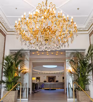 Luksuriøs hotellobby med en storslått lysekrone, marmorvegger og elegant innredning. To store potteplanter rammer inn en passasje til en sittegruppe som er synlig i bakgrunnen. Varm belysning forsterker den overdådige atmosfæren.