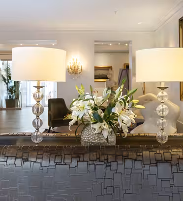 Elegant hotellobby med mønstret sofa, to lamper med krystallsokler og en bukett med hvite liljer. Myk belysning fra vegglamper og en utsmykket ramme på veggen bidrar til den sofistikerte stemningen.