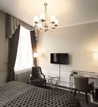 Et elegant hotellrom har en seng med et blomstermønstret sengeteppe, et vindu med kraftige gardiner, en flatskjerm-TV på veggen, en moderne lysekrone, et skrivebord med en lampe og en koselig lenestol. Innredningen er nøytral og sofistikert.