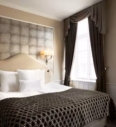Elegant hotellrom med stor seng, mønstret teppe og flere puter. Veggen har sconces med myk belysning og teksturert panel bak sengegavlen. Høye draperte gardiner rammer inn et vindu til høyre og slipper inn naturlig lys.