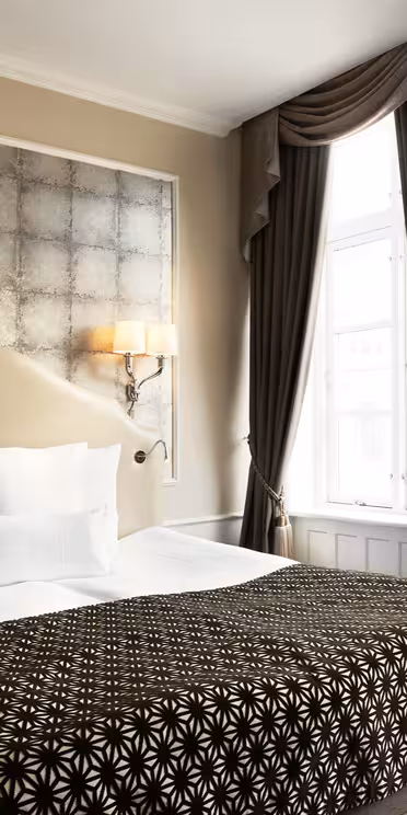 Elegant hotellrom med stor seng, mønstret teppe og flere puter. Veggen har sconces med myk belysning og teksturert panel bak sengegavlen. Høye draperte gardiner rammer inn et vindu til høyre og slipper inn naturlig lys.