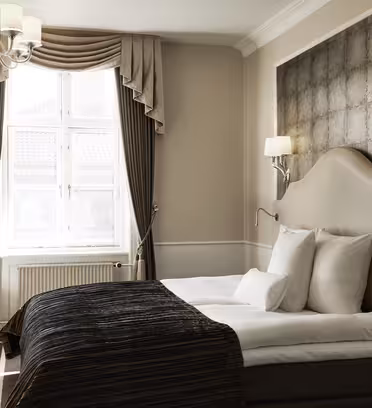 Et stilig hotellrom med stor seng, elegant hodegjerde og mørkt sengetøy. Lamper flankerer sengen, og et vindu med kraftige gardiner slipper inn naturlig lys. En lysekrone henger fra taket, og et skrivebord med glassplate er i hjørnet.