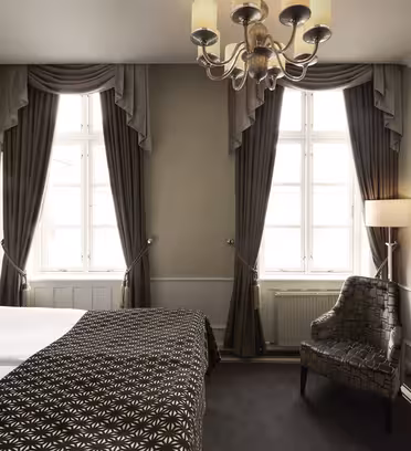 Elegant hotellrom med dobbeltseng og mønstret dyne. To høye vinduer med mørke gardiner, en lysekrone og en mønstret lenestol. En flatskjerm-TV er montert på veggen over et sidebord i glass. Nøytrale farger dominerer innredningen.