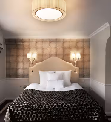 Elegant hotellrom med en stor seng med polstret sengegavl og mønstret sengeteppe. To veggmonterte lamper lyser opp den strukturerte veggen bak. Et stort vindu med gardiner er til venstre, og et lite sidebord med glassplate står ved siden av sengen.