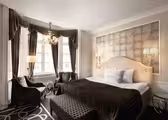 Et luksuriøst hotellrom har en stor seng med mønstret hodegjerde, elegante gardiner og en lysekrone. Det er en sittegruppe med to lenestoler og et lite bord ved vinduene. Nøytrale toner og dekorativ belysning gir raffinement til rommet.