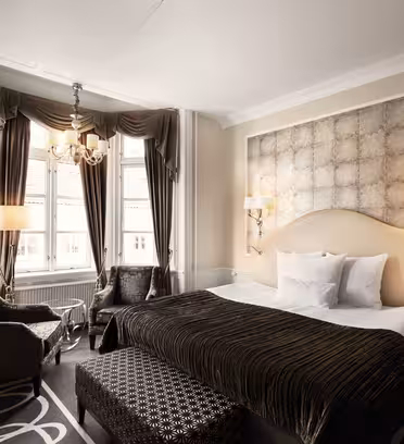 Et luksuriøst hotellrom har en stor seng med mønstret hodegjerde, elegante gardiner og en lysekrone. Det er en sittegruppe med to lenestoler og et lite bord ved vinduene. Nøytrale toner og dekorativ belysning gir raffinement til rommet.