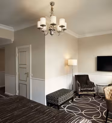 Elegant hotellrom med beige vegger, en stor flatskjerm-TV, lenestoler og en mønstret ottoman. Det er en lampe ved vinduet og en lysekrone over hodet. Gardiner dekker delvis et vindu ved siden av et skrivebord og en stol. En seng er delvis synlig.