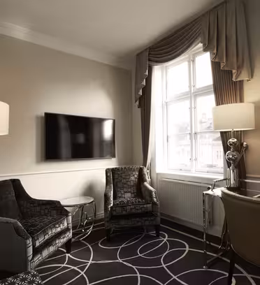 Et moderne hotellrom med to mørkmønstrede lenestoler, et lite salongbord i glass, en flatskjerm-TV på veggen og et skrivebord med en stol. Et stort vindu er dekket med beige gardiner, og to høye lamper lyser opp rommet.