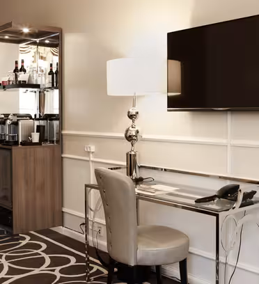 Hotellromshjørne med skrivebord i glass, stol og en elegant bordlampe. En flatskjerm-TV er montert på den beige veggen. Til venstre er det en minibar med glasshyller med flasker og en kaffestasjon, ved siden av et skap. Geometrisk mønstret teppe.