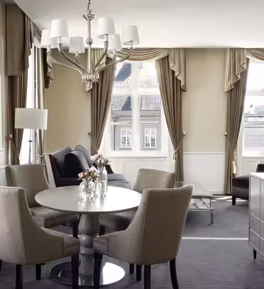 Elegant hotellsuite med rundt spisebord, fire beige stoler og en mørkegrå sofa. Store vinduer med beige gardiner gir rikelig med naturlig lys. En lysekrone henger over, og blomster pynter på bordet. Nøytrale toner dominerer innredningen.