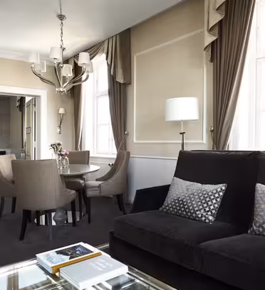 Elegant hotellsuite med stue med svart sofa, mønstrede puter og salongbord i glass med bøker. Et rundt spisebord med fire stoler er nær inngangen til et soverom med seng og lysekrone. Nøytrale toner og store vinduer forsterker lyset.