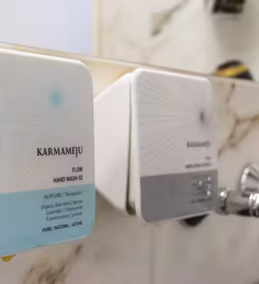 To firkantede dispenserflasker montert på en marmorvegg, merket "Karmameju" med en tekst "Flow Hand Wash 02." Kraner er synlige under dispenserne. Innredningen er moderne med en luksuriøs følelse.