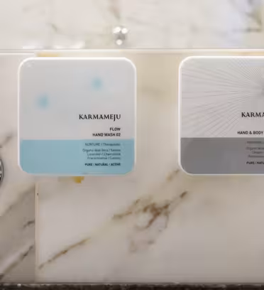 Veggmonterte dispensere merket "FLOW" og "CHILL" av Karmameju på en marmorvegg, flankert av kromkraner. Dispenserne inneholder henholdsvis håndvask og hånd- og bodylotion.