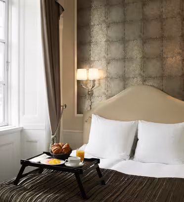 Et koselig hotellrom har en pent oppredd seng med en beige hodegjerde mot en teksturert vegg. Et frokostbrett med croissanter, frukt og appelsinjuice ligger på sengen. To vegglamper lyser opp rommet, og et vindu med brune gardiner er i nærheten.