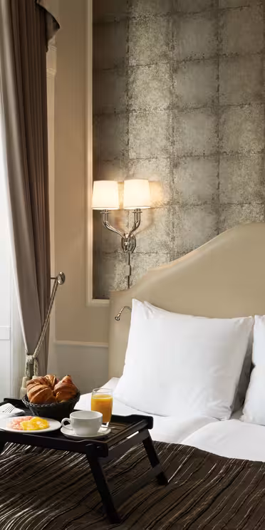 Et koselig hotellrom har en pent oppredd seng med en beige hodegjerde mot en teksturert vegg. Et frokostbrett med croissanter, frukt og appelsinjuice ligger på sengen. To vegglamper lyser opp rommet, og et vindu med brune gardiner er i nærheten.