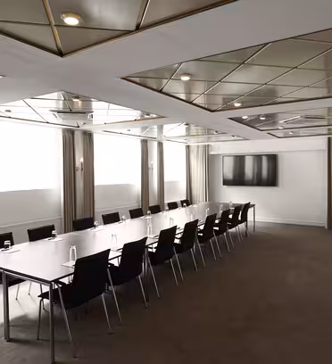 Et moderne konferanserom med et langt rektangulært bord omgitt av svarte stoler. Taket har geometriske mønstre med innfelte lys. En stor flatskjerm-TV er montert på veggen, og to store vinduer med beige gardiner gir naturlig lys.