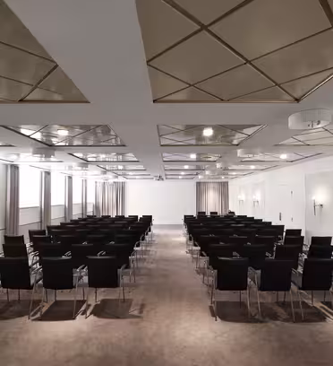 Et moderne konferanserom med rader med tomme svarte stoler vendt mot en hvit projeksjonsskjerm. Rommet har geometrisk takdesign, dempet belysning og beige tepper. Store vinduer med gardiner langs den ene siden av rommet.
