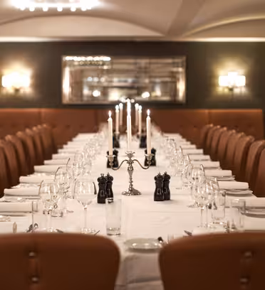 Et langt, elegant dekket spisebord med hvite duker strekker seg gjennom en varmt opplyst restaurant. Flere lysestaker med tente stearinlys, glass og sorte salt- og pepperbøsser pryder bordet. Skinnstoler på begge sider.