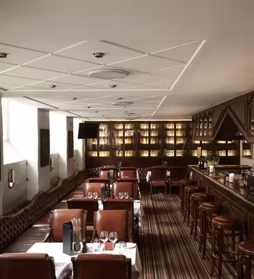 En svakt opplyst, elegant bar med skinnseter, en polert trebenk og hyller foret med flasker. Bord dekkes med hvite duker og vinglass. Varm belysning skaper en koselig atmosfære.