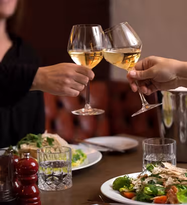 To personer skåler med glass hvitvin over et bord dekket med salatretter, et stearinlys og en pepperkvern. En vinflaske i en isbøtte er i bakgrunnen. Scenen fanger en koselig spiseatmosfære.