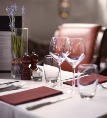 Elegant restaurantbordsett for to, med hvit duk, røde servietter, vin- og vannglass, bestikk, salt- og pepperbøsse, og en liten vase med blomster. To røde stoler er plassert ved bordet. Myk belysning fyller scenen.
