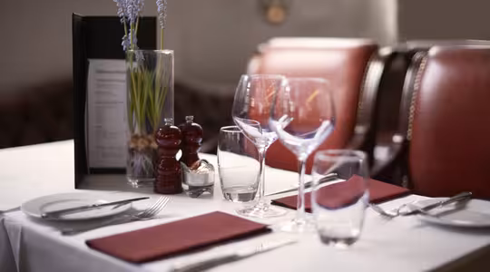 Elegant restaurantbordsett for to, med hvit duk, røde servietter, vin- og vannglass, bestikk, salt- og pepperbøsse, og en liten vase med blomster. To røde stoler er plassert ved bordet. Myk belysning fyller scenen.