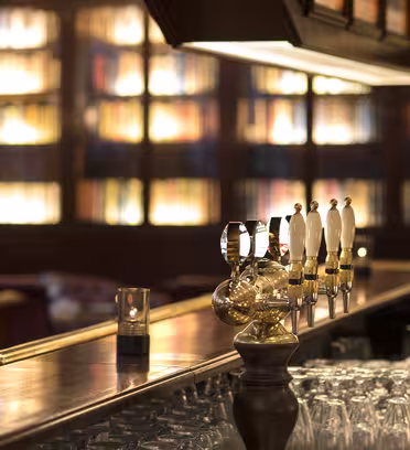 En koselig bar med benkeplater i tre og flere ølkraner. Hyller med glødende bøker fyller den mykt opplyste bakgrunnen. Rader med glass er pent ordnet under disken, og et enkelt lys flimrer på baren.